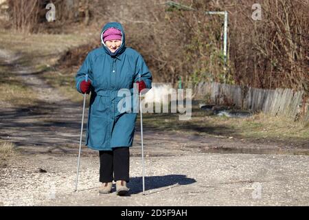 Femme âgée marchant avec des bâtons sur la route rurale, des exercices sportifs pour une colonne vertébrale et des articulations saines. Marche nordique par temps froid, thérapie pour la santé Banque D'Images