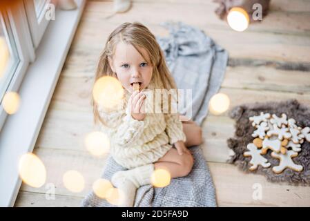 Une petite fille dans un chandail blanc tricoté boit du chocolat chaud, du cacao. Le bébé regarde par la fenêtre, attend le miracle du nouvel an le Père Noël, mange Banque D'Images