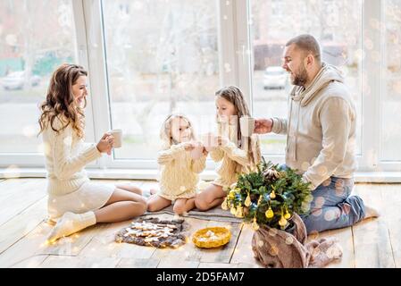 Une famille heureuse, avec des chandails blancs, boit du cacao près de la fenêtre panoramique. Les parents et les filles tiennent des tasses dans leurs mains, sur une assiette sont du pain d'épice Banque D'Images