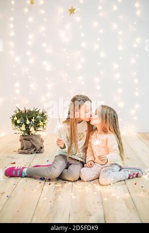 Heureux deux amis scincheurs de lumière par un petit arbre de Noël dans un pot sur un fond de bokeh flou de guirlandes jaunes, lumineuses. Les sœurs a Banque D'Images