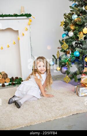 Portrait rapproché d'une petite fille blonde dans une robe blanche près d'un arbre de Noël avec une guirlande lumineuse. Vacances du nouvel an près de la cheminée à la maison. Banque D'Images