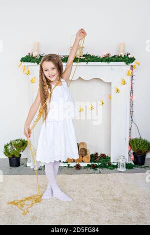 Portrait rapproché d'une petite fille blonde dans une robe blanche près d'un arbre de Noël avec une guirlande lumineuse. Vacances du nouvel an près de la cheminée à la maison. Banque D'Images