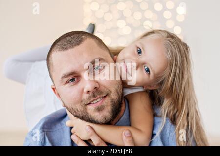 Un portrait en gros plan d'un père heureux et d'une petite fille sur un fond blanc de bokeh jaune. Un jeune homme avec une barbe joue et épouse sa fille. Banque D'Images