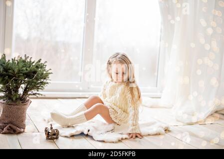 Petite fille heureuse dans une robe de chandail blanc tricoté, s'amusant par la fenêtre panoramique, dansant, décorant un arbre de Noël dans un pot avec une guirlande Banque D'Images