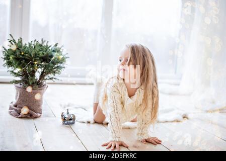 Petite fille heureuse dans une robe de chandail blanc tricoté, s'amusant par la fenêtre panoramique, dansant, décorant un arbre de Noël dans un pot avec une guirlande Banque D'Images