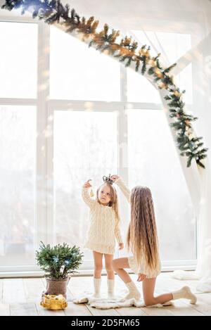 Deux amies de la sœur blonde en chandails blancs tricotés Et les chaussettes jouent par la fenêtre panoramique avec Noël arbres en prévision Banque D'Images