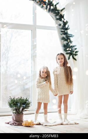 Deux amies de la sœur blonde en chandails blancs tricotés Et les chaussettes jouent par la fenêtre panoramique avec Noël arbres en prévision Banque D'Images