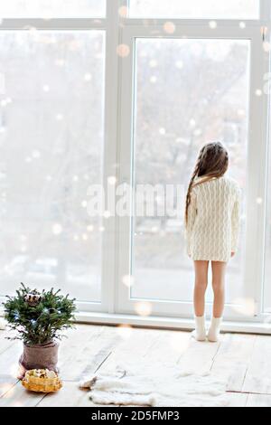 Petite fille heureuse dans une robe de chandail blanc tricoté, regarde dans la fenêtre panoramique, danse, décore un arbre de Noël dans un pot avec une guirlande de lig Banque D'Images