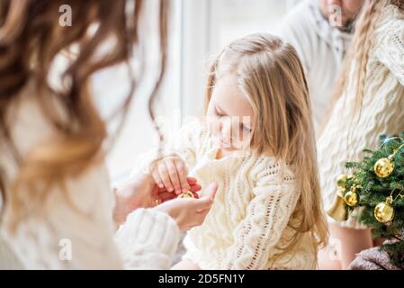 Une mère et une fille blonde en chandails tricotés blancs décorent un petit arbre de Noël avec des jouets dorés. Une jeune femme montre entre les mains la fille nouvel an Banque D'Images