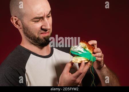 Le concept de restauration rapide et d'écologie. Un homme chauve avec une barbe ressemble déguisement à la garniture en plastique d'un hamburger. Arrière-plan Bourgogne. Gros plan Banque D'Images