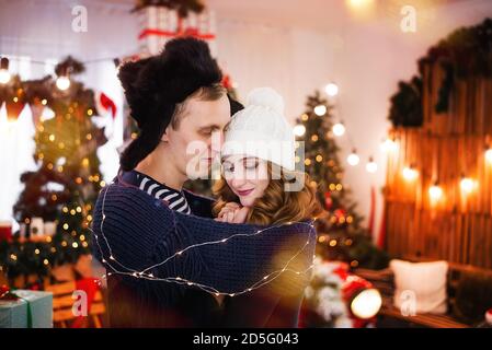 Un portrait en gros plan d'un jeune couple amoureux sur fond d'arbres de Noël avec guirlandes. Un jeune homme dans un chapeau avec des rabats chauffe Banque D'Images