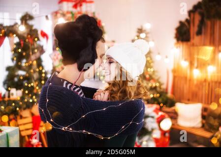 Un portrait en gros plan d'un jeune couple amoureux sur fond d'arbres de Noël avec guirlandes. Un jeune homme dans un chapeau avec des rabats chauffe Banque D'Images