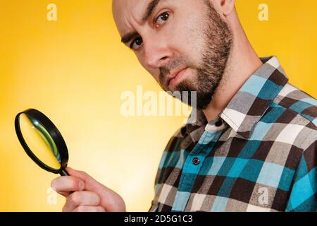 Le concept de la recherche et de la résolution des problèmes. Un homme chauve avec une barbe, habillé dans une chemise à carreaux bleue, tenant une loupe et regardant attentivement. Banque D'Images