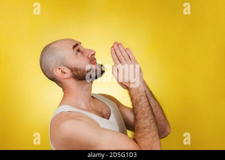 Les gens et les émotions. Un homme chauve, barbu dans un t-shirt blanc a humblement clasé les mains dans la prière ou la demande. Arrière-plan jaune. Copier l'espace. Banque D'Images