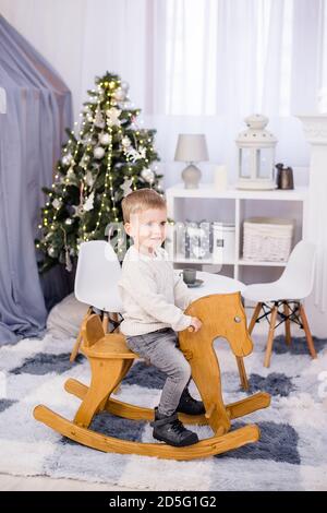 Joyeux petit garçon blond dans un chandail blanc tricoté et un jean se déplace sur un cheval à bascule en bois dans la chambre des enfants À côté d'un arbre de Noël avec guirlandes Banque D'Images