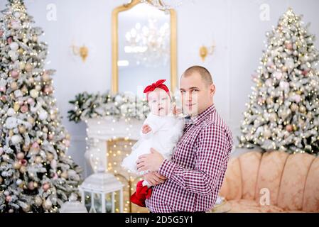 Un jeune homme tient un bébé dans ses bras. Le père joue avec une fille dans une robe blanche, l'embrasse, embrasse contre le fond des arbres de Noël festifs Banque D'Images