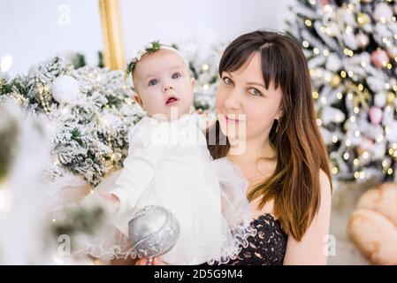 Une jeune belle femme dans une robe de soirée luxuriante tient, embrasse, embrasse un petit bébé dans une robe blanche. Sur fond d'arbres enneigés Banque D'Images