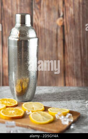 Tranches d'orange et de citron recouvertes de glace sur une planche de bois sur une surface grise, avec un shaker à cocktail derrière Banque D'Images