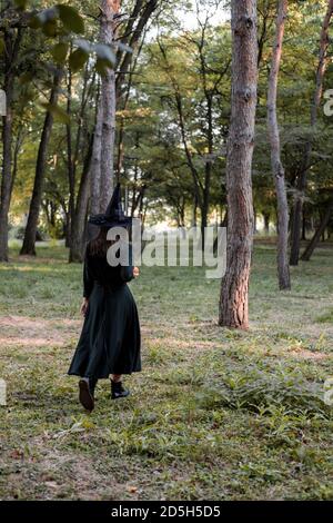 Une jeune femme en robe noire et chapeau de sorcière tient une citrouille dans ses mains. Costume de fête d'Halloween. Forêt, parc avec arbres d'automne Banque D'Images