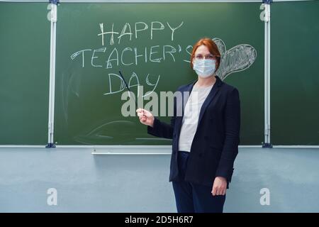 Une femme dans un masque médical et avec un pointeur près du tableau noir avec le texte « Happy Teacher's Day ». Concept des problèmes d'apprentissage pendant la quarantaine Banque D'Images