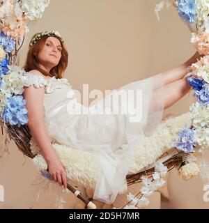 Une femme souriante est assise avec une flûte à bloc sur une balançoire en fleurs. Portrait de studio de la jolie femme flûtiste en robe rose Banque D'Images