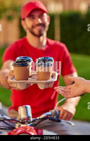 Un jeune messager souriant donne des tasses de café au client dans une rue ensoleillée. Mise au point sélective. Concept de livraison de nourriture Banque D'Images