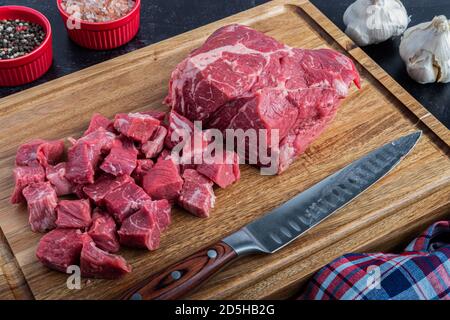 Un rôti de bœuf désossé est coupé en cubed pour un ragoût sur une planche à découper avec du sel de mer et des grains de poivre à un couteau à découper Banque D'Images