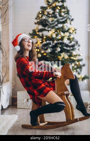 Noël, nouvel an. Jolie femme dans la chemise et les chaussettes ont l'amusement équitation sur le jouet cheval balançoire en bois, arbre de nouvel an derrière Banque D'Images