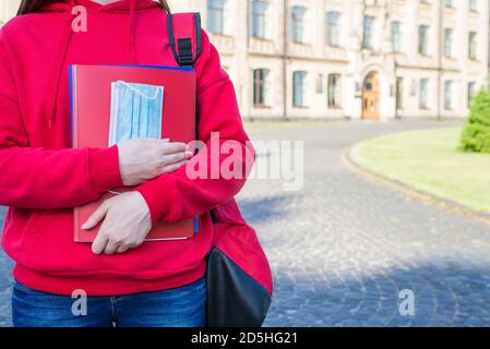 Premier jour à l'école après le concept d'université. Photo courte en gros plan d'une jeune fille dans un chandail rouge décontracté tenant des livres et un masque médical debout près d Banque D'Images