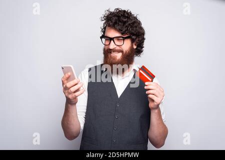 Un jeune barbu tient une voiture de crédit et un téléphone portant ses lunettes est souriant et regarde dans le téléphone. Banque D'Images