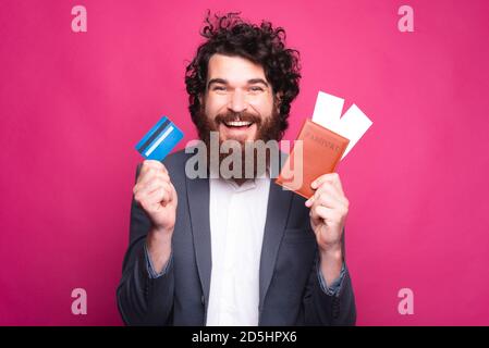 Un homme à la barbe est heureux de tenir une carte de crédit et un passeport avec deux billets dans lui près d'un mur rose. Banque D'Images