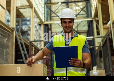 Jeune ouvrier indien d'entrepôt industriel d'usine travaillant dans l'industrie logistique intérieure. Souriant heureux tenant une planchette à pince pour vérifier la commande de marchandise en magasin Banque D'Images