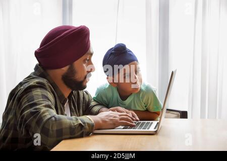 UN ENFANT SIKH REGARDANT ATTENTIVEMENT LE TRAVAIL EFFECTUÉ PAR PÈRE SUR UN ORDINATEUR PORTABLE Banque D'Images