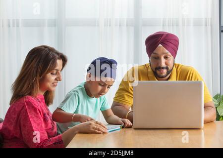 UN ENFANT SIKH LISANT AVEC SA MÈRE ALORS QUE SON PÈRE TRAVAILLE SUR SON ORDINATEUR PORTABLE Banque D'Images