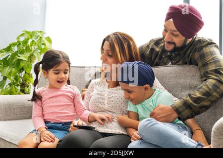 UN ENFANT SIKH REGARDANT LE TÉLÉPHONE MOBILE AVEC PÈRE ET LA MÈRE REGARDE AVEC JOIE SA SŒUR QUI PARLE Banque D'Images