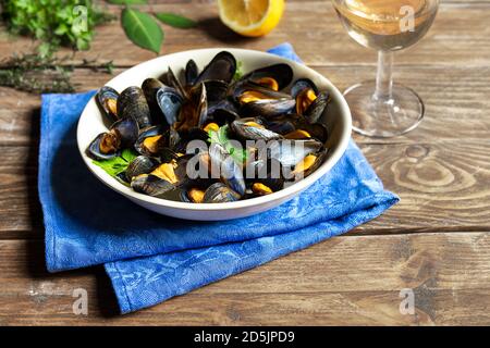 Délicieuses moules fraîches cuites à la vapeur dans une sauce au vin blanc dans une assiette blanche, un citron, du persil et un verre de vin sur une table en bois Banque D'Images