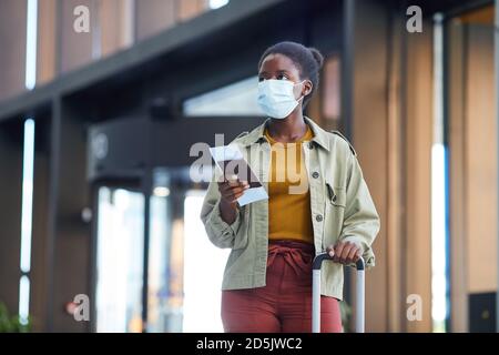 Jeune femme africaine dans un masque de protection avec des bagages tenant des billets en allant le long de l'aéroport Banque D'Images