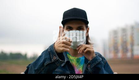 Beau jeune homme en chapeau, masque chirurgical, médical et montrer le gros doigt sur la rue dans la ville par temps nuageux. Coronavirus, protection Covid-19. Bannière Banque D'Images