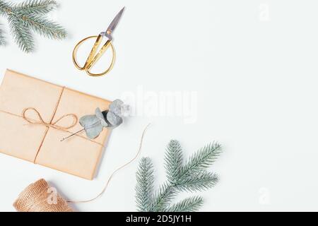 Emballage cadeau maison de Noël avec boîte en papier artisanal, ciseaux dorés, branches de sapin et d'eucalyptus sur fond blanc avec espace de copie. Banque D'Images