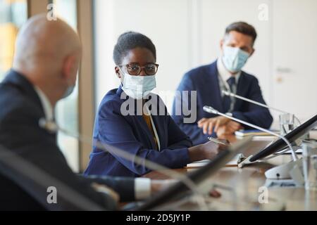 Portrait de la jeune femme d'affaires africaine dans le masque de protection regardant appareil photo lors d'une réunion avec ses collègues Banque D'Images