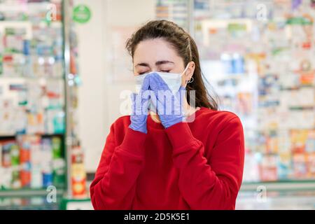 Une jeune femme dans un masque médical et des gants éternue, couvrant son visage. En arrière-plan, les fenêtres d'achat des produits pharmaceutiques. Concept de la maladie, v Banque D'Images