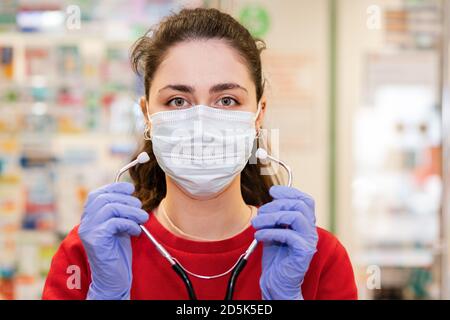 Portrait d'une femme dans un masque médical, des gants et des vêtements rouges, tenant un stéthoscope près de son visage. Fenêtre pharmacie en arrière-plan. Concept de Banque D'Images