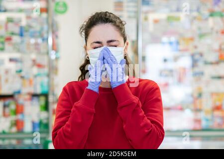 Une jeune femme dans un masque médical et des gants éternue, couvrant son visage. En arrière-plan, les fenêtres d'achat des produits pharmaceutiques. Concept de la maladie, v Banque D'Images