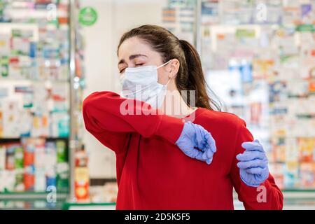 Une jeune femme dans un masque médical et des gants tousse, couvrant son visage avec le creux de son coude. En arrière-plan, les fenêtres de magasin de la pharmacie prod Banque D'Images