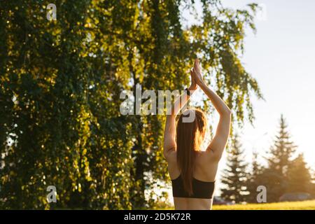 Vue arrière d'une jeune femme méconnue assise sur un tapis de yoga en position lotus et levant les mains vers le haut. Banque D'Images