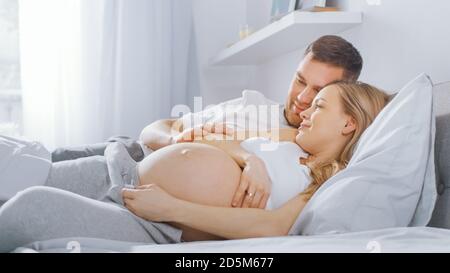 Un jeune couple heureux se câliner dans le lit, la jeune femme est enceinte et affectueuse touche son mari et la caresse tendresse de son ventre. Mise au point de l'appareil photo activée Banque D'Images
