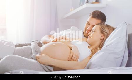 Un jeune couple heureux se câliner dans le lit, la jeune femme est enceinte et affectueuse touche son mari et la caresse tendresse de son ventre. Mise au point de l'appareil photo activée Banque D'Images