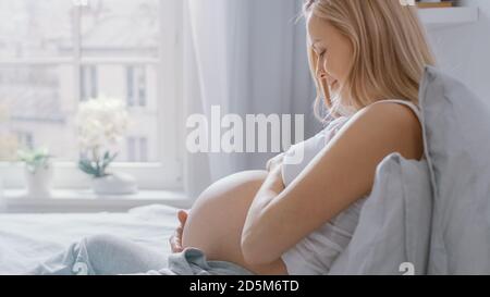 Bonne jeune femme enceinte posée au lit le matin, elle touche et caresse sa Belly tendly, aimant son futur bébé et chaque moment d'elle Banque D'Images