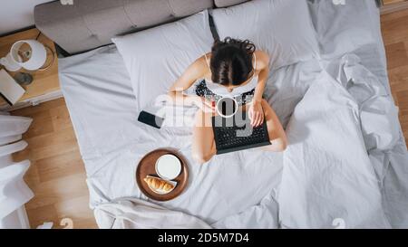 Efficace jeune fille millénaire assise sur un lit le matin, utilise un ordinateur portable et mange des croissants et des boissons café pour le petit déjeuner. Capote abaissée Banque D'Images