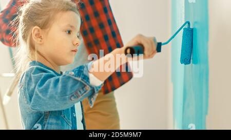 Adorable petite fille en Jean manteau est la peinture d'un mur. Elle peint avec le rouleau qui est couvert de peinture bleu clair. Le père peint avec sa fille Banque D'Images